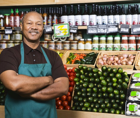 Smiling Jamaican shop owner wearing a green apron stands confidently in front of neatly arranged fresh produce and bottled goods, representing empowered MSMEs benefiting from accessible business loans through WILCO Finance.