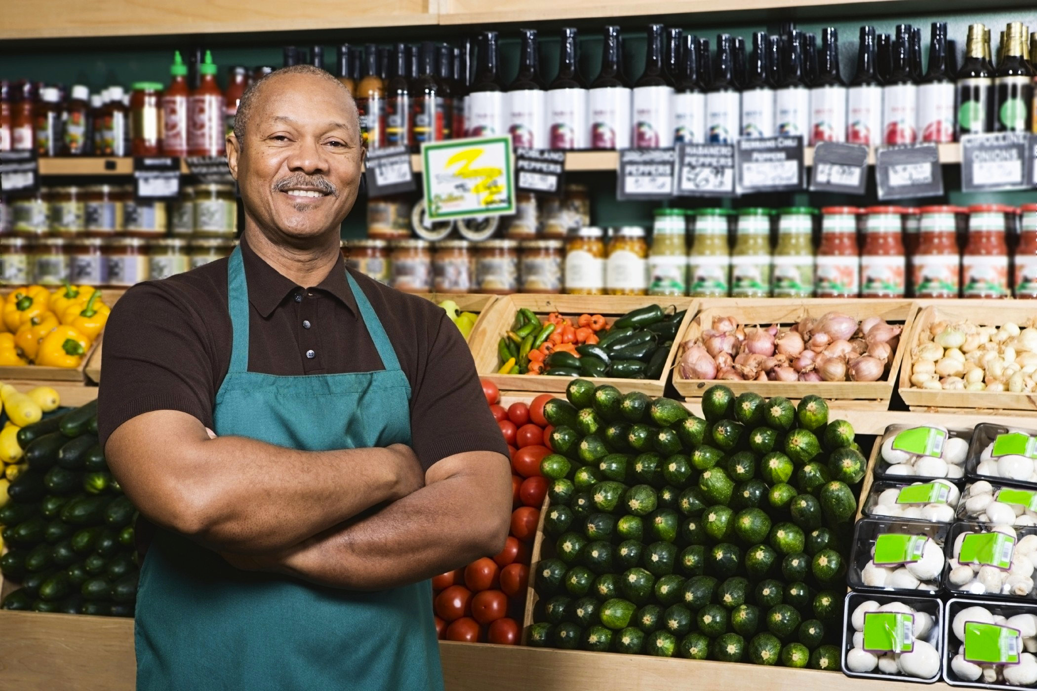 Smiling Jamaican shop owner wearing a green apron stands confidently in front of neatly arranged fresh produce and bottled goods, representing empowered MSMEs benefiting from accessible business loans through WILCO Finance.
