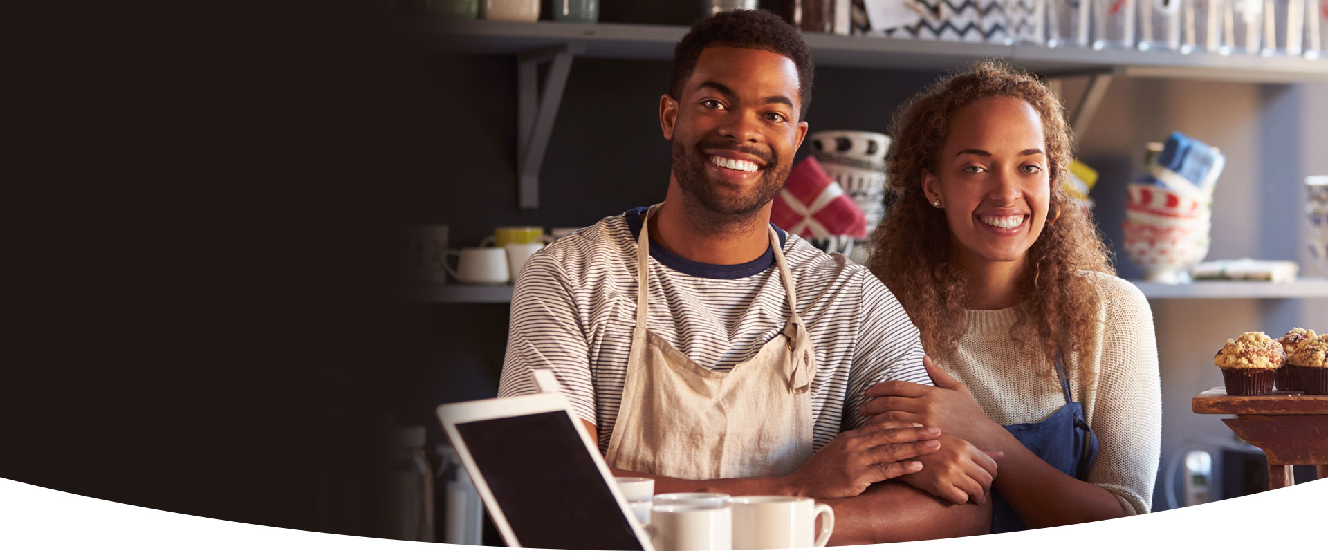 A smiling young Jamaican couple wearing aprons sits together in their cozy café, surrounded by mugs, pastries, and warm light — representing small business owners supported by WILCO Finance.