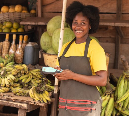 Smiling young female Jamaican fruit vendor in a yellow shirt and apron stands beside bunches of bananas and tropical produce, representing small business owners empowered by WILCO Finance MSME loans.