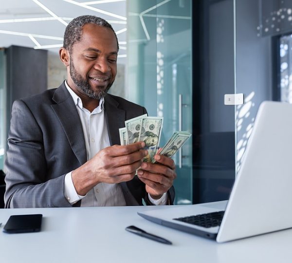 Smiling businessman in a modern office counts U.S. hundred‑dollar bills beside a laptop, symbolizing improved cash flow and financial growth made possible through WILCO Finance’s small business loan solutions in Jamaica.