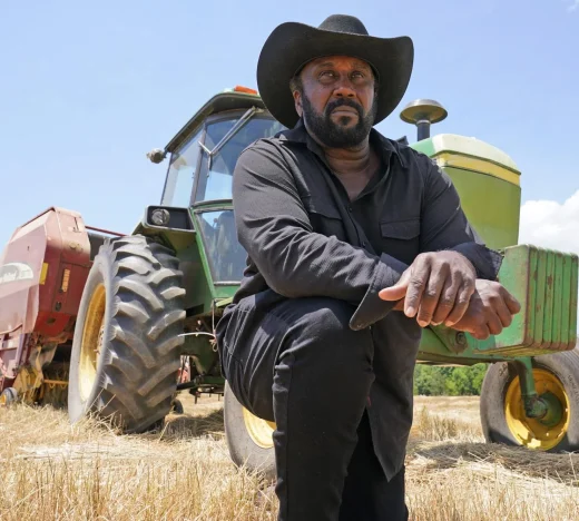 Confident Jamaican farmer wearing a black hat kneels beside a green tractor in a harvested field, representing agricultural entrepreneurs expanding operations with MSME financing.