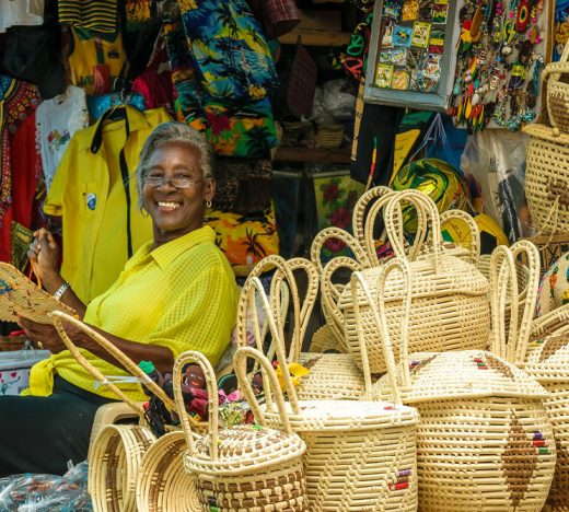 Smiling Jamaican artisan in a yellow shirt sits among handmade wicker baskets and colorful merchandise, showcasing local craftsmanship supported by WILCO Finance small business loans.